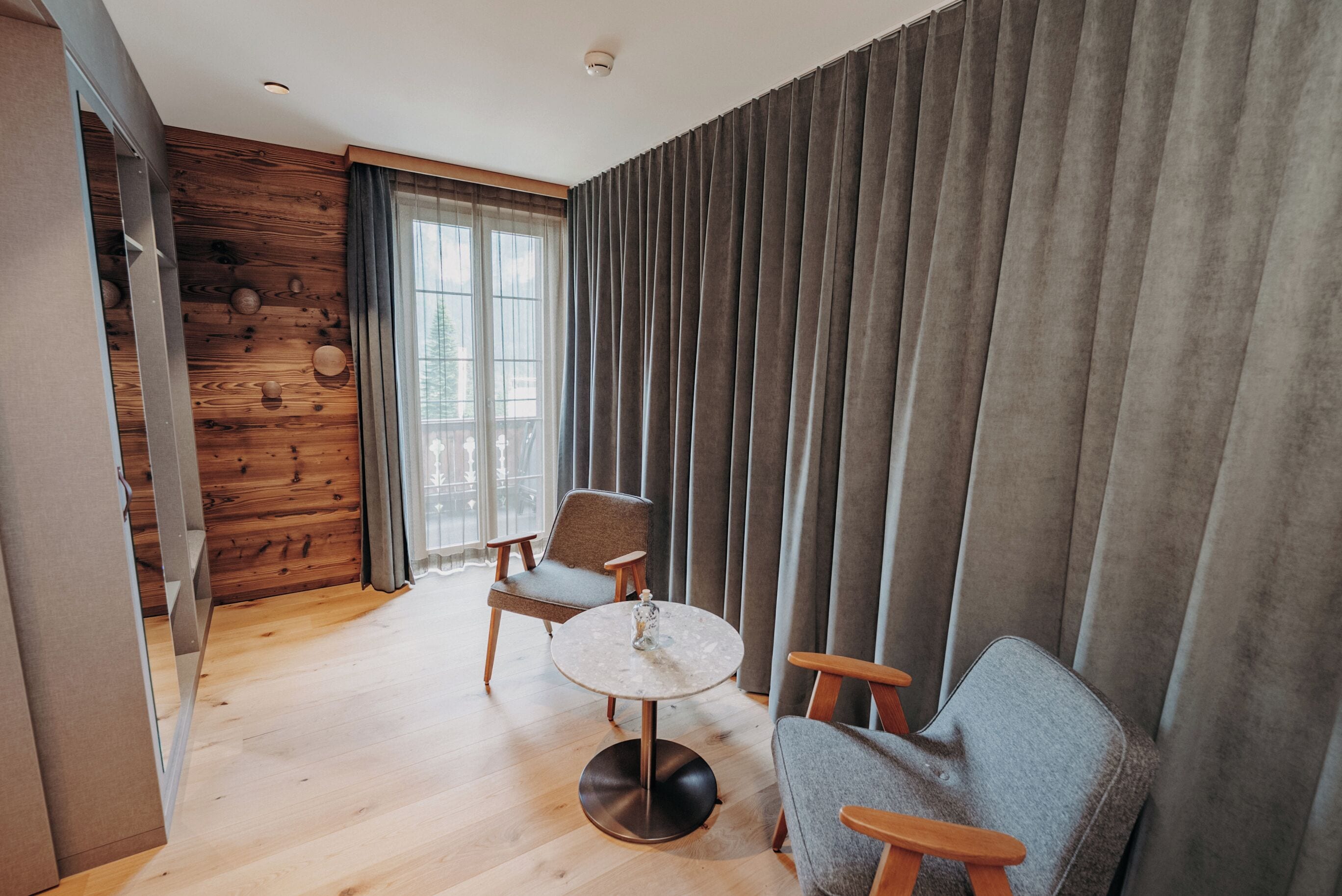 A modern room with wooden panel walls, a round table, two chairs, and a large window with curtains.