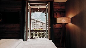 A hotel room with a bed, a wooden headboard, a nightstand, a lamp, and a view of a mountain and a building through an open door. - Parkhotel Schoenegg (Grindelwald)
