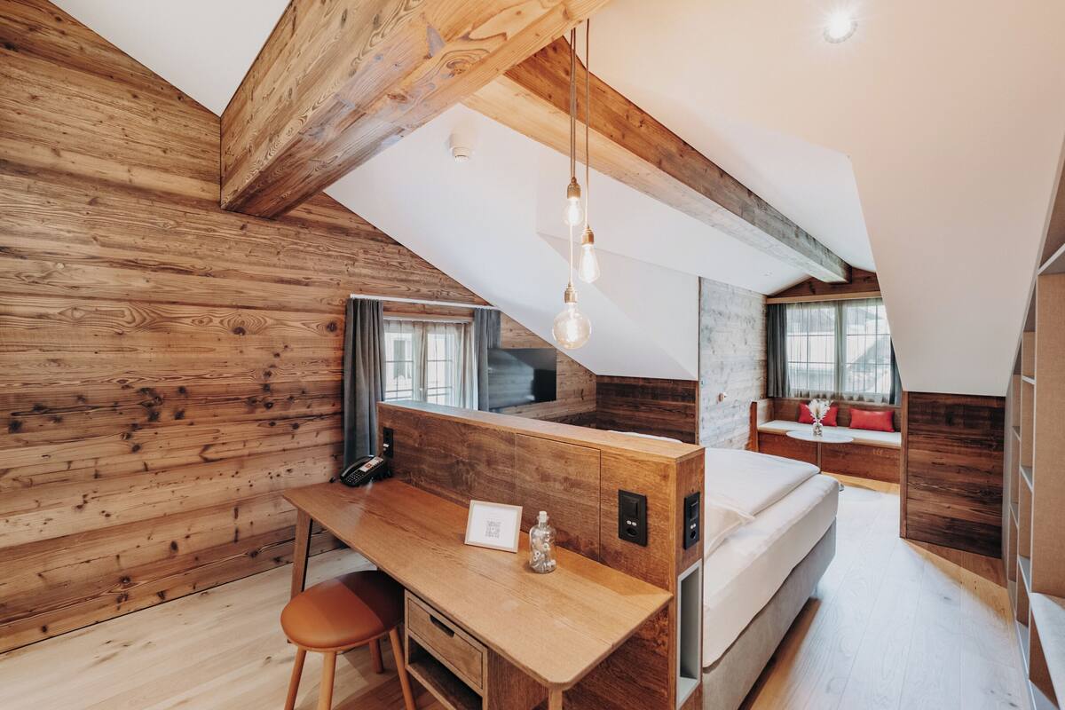 A loft-style bedroom with a wooden ceiling, a desk with a chair, a bed, and a window with curtains.
