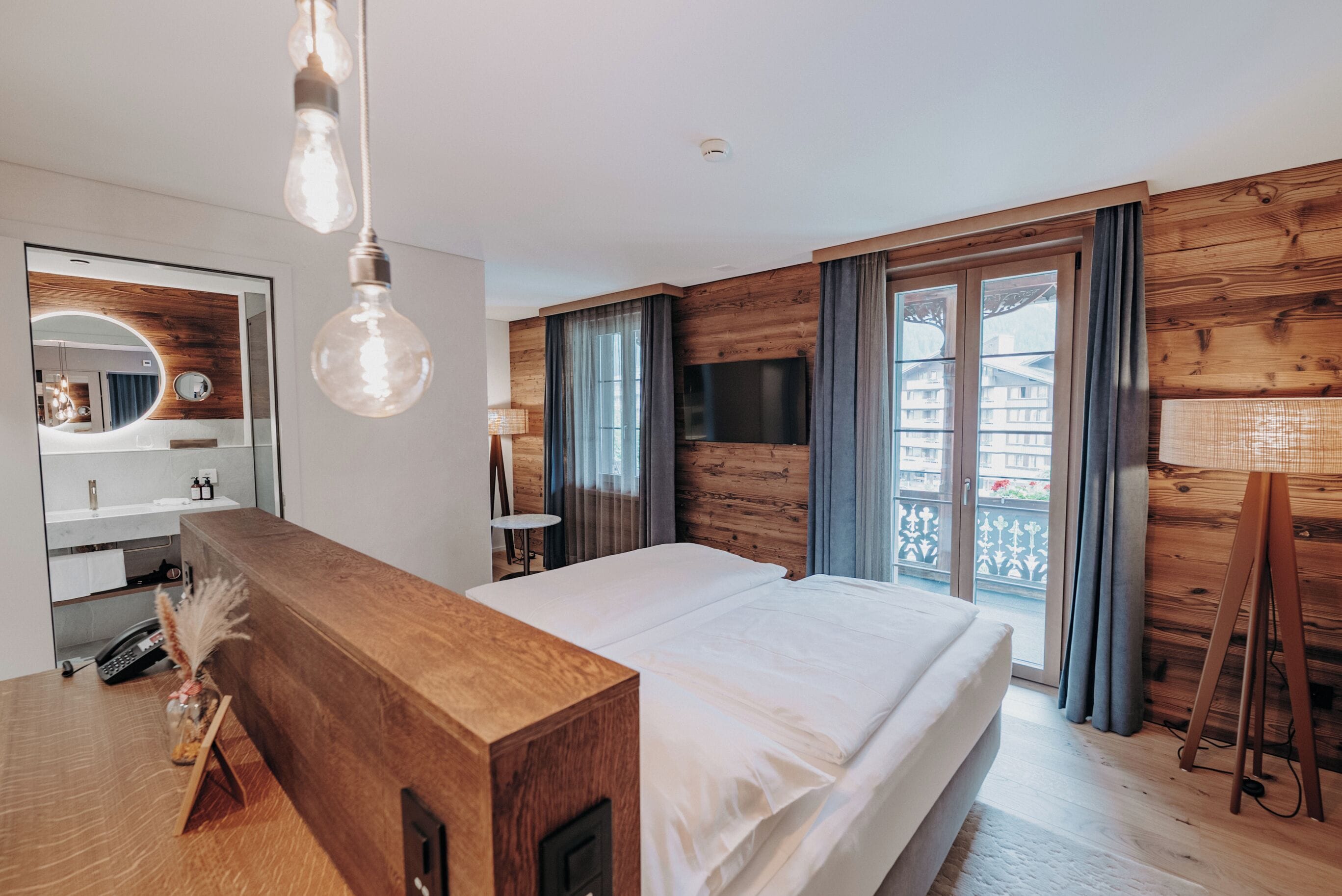 A modern bedroom with a wooden headboard, a large bed, a TV, and a balcony with curtains.