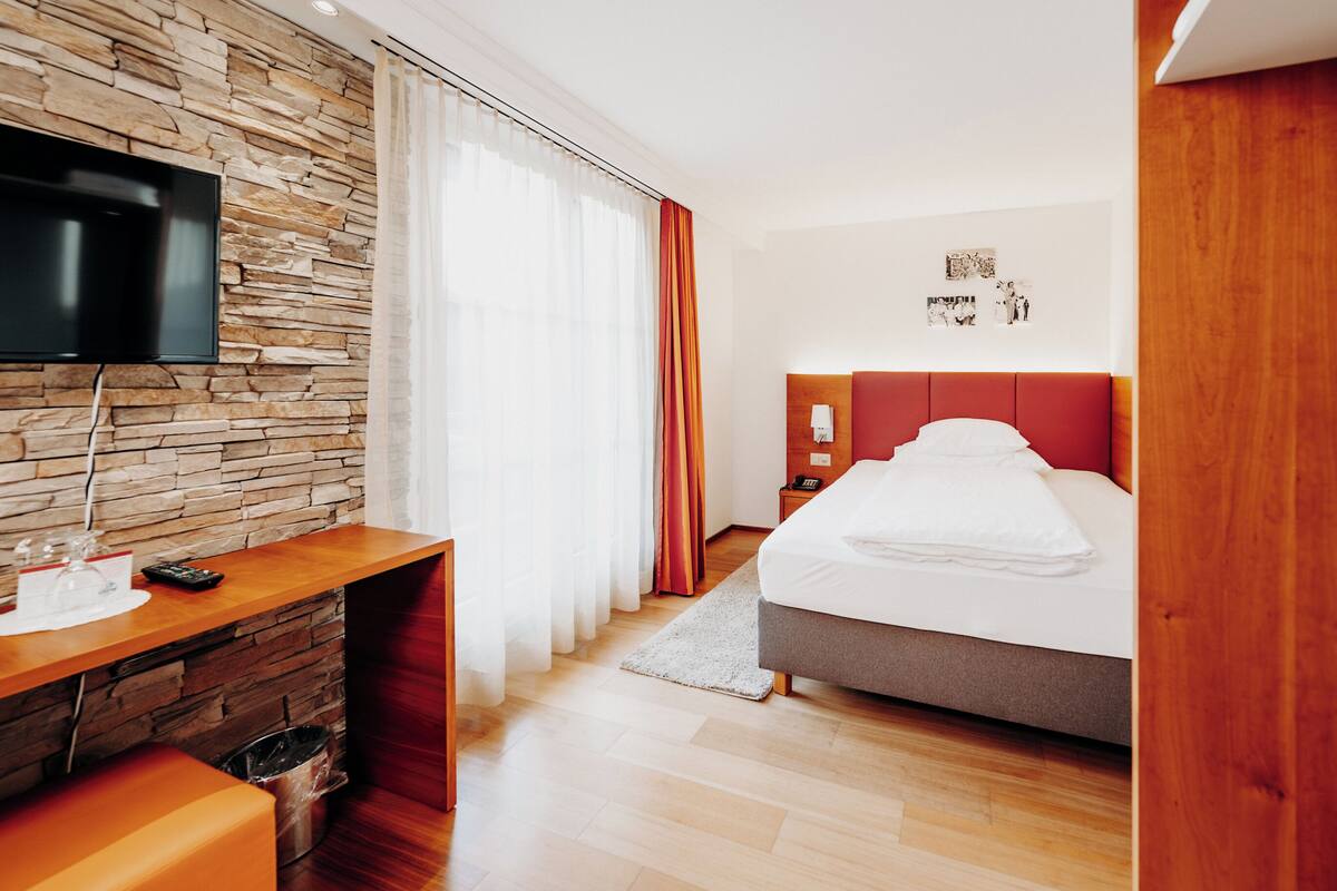 A hotel room with a stone wall, a flat-screen TV, a bed with a red headboard, a desk with a computer, and a small trash bin.