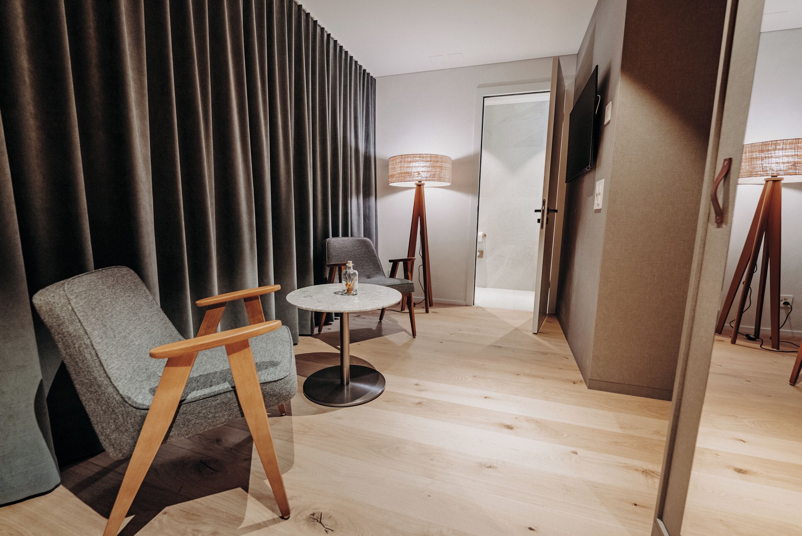 Una habitación de hotel moderna con suelo de madera, un sillón gris, una pequeña mesa redonda, una lámpara de pie y un televisor montado en la pared.