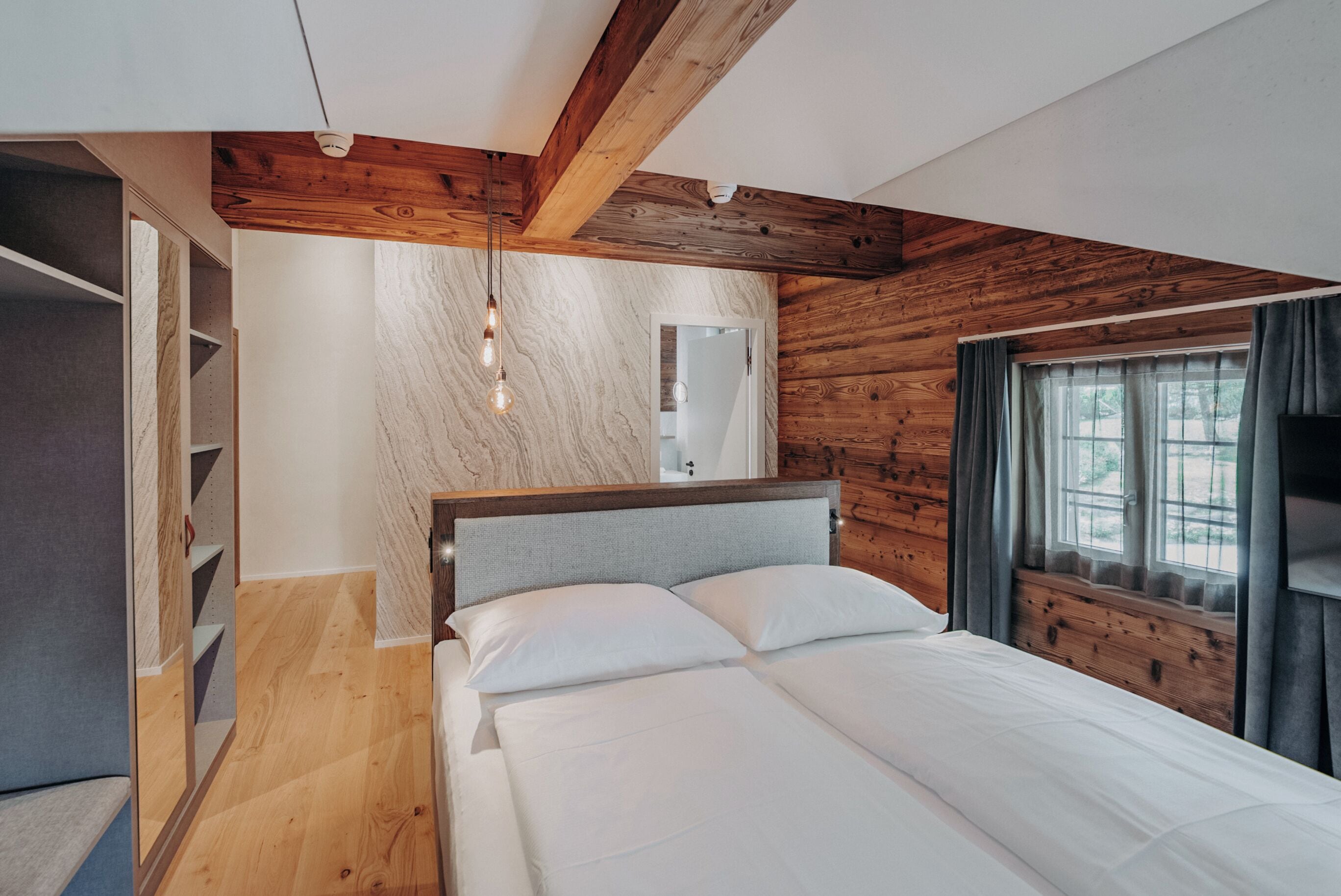 A bedroom with a wooden ceiling, a bed with white linens, a built-in wardrobe, and a window with curtains.