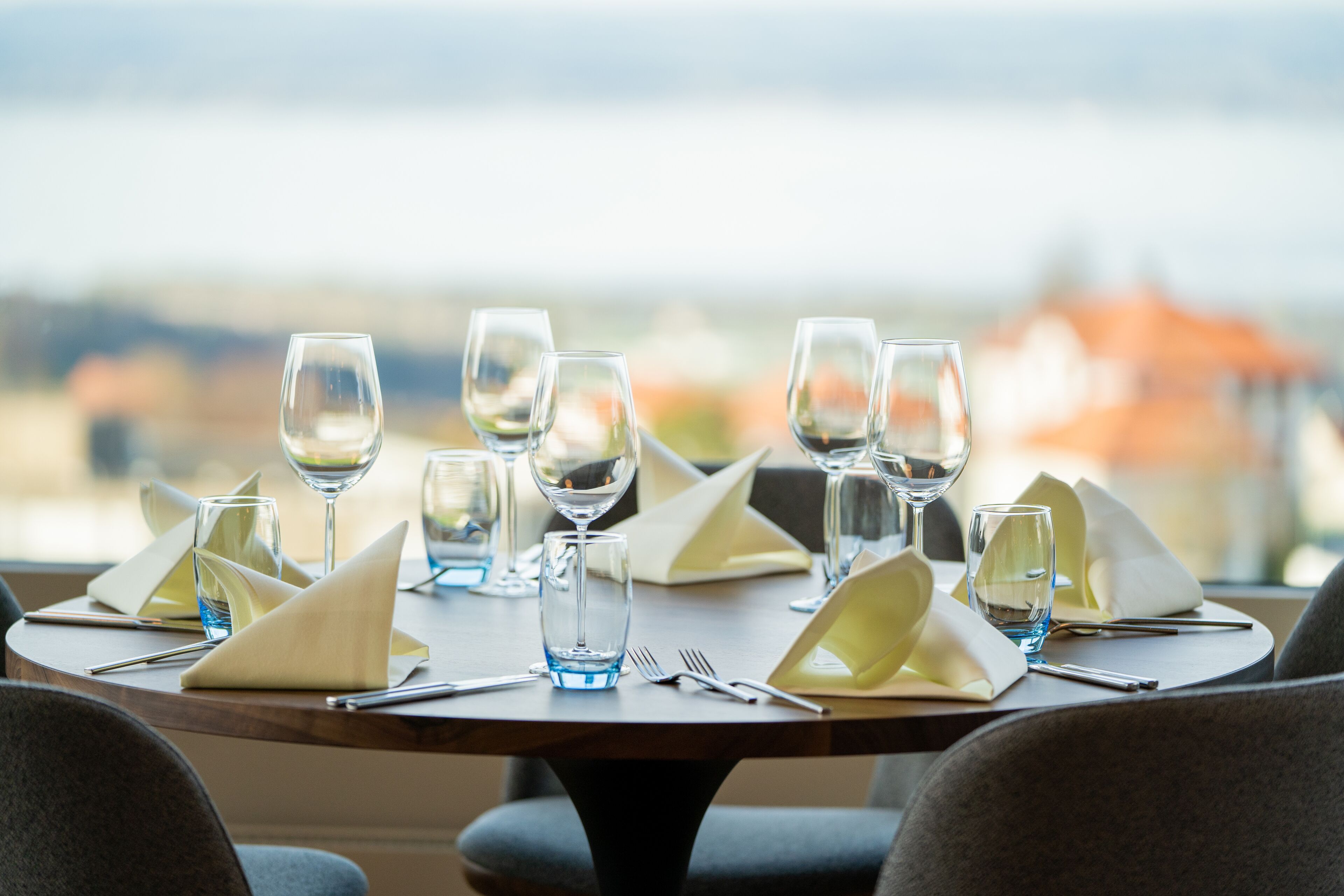 Una mesa de comedor lista para una comida, con varias copas de vino, vasos de agua y cubiertos, con vistas a edificios al fondo.