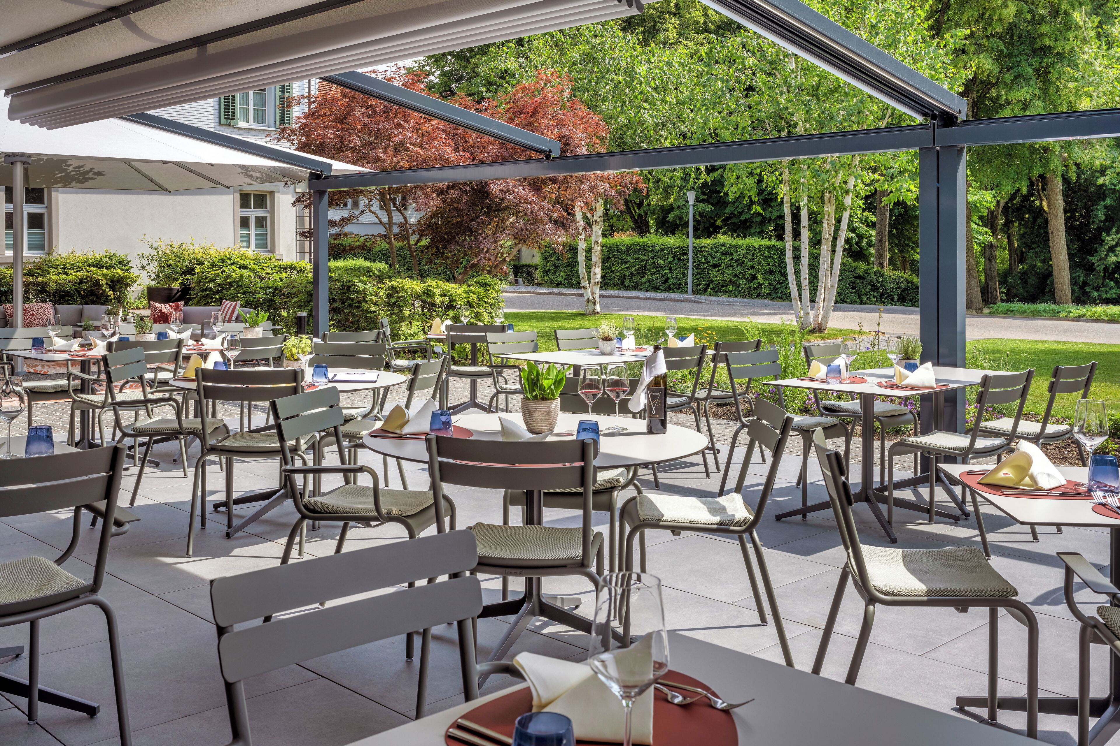 Eine Terrasse im Freien mit mehreren Tischen und Stühlen, umgeben von Grünpflanzen und einem Gebäude im Hintergrund.