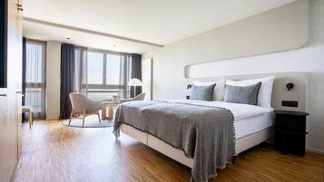 A modern hotel room with a large bed, a chair, a small table, and a view of the outdoors.