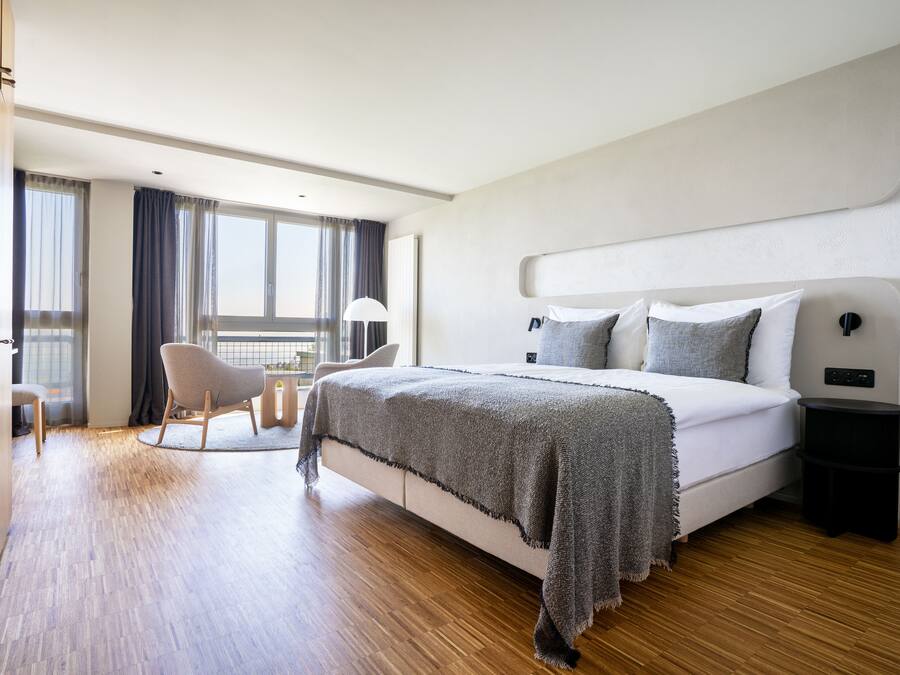 A modern hotel room with a large bed, a chair, a small table, and a view of the outdoors.