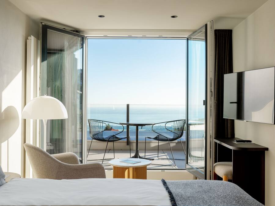 A modern hotel room with a bed, a desk, a TV, and a balcony with a view of the sea.