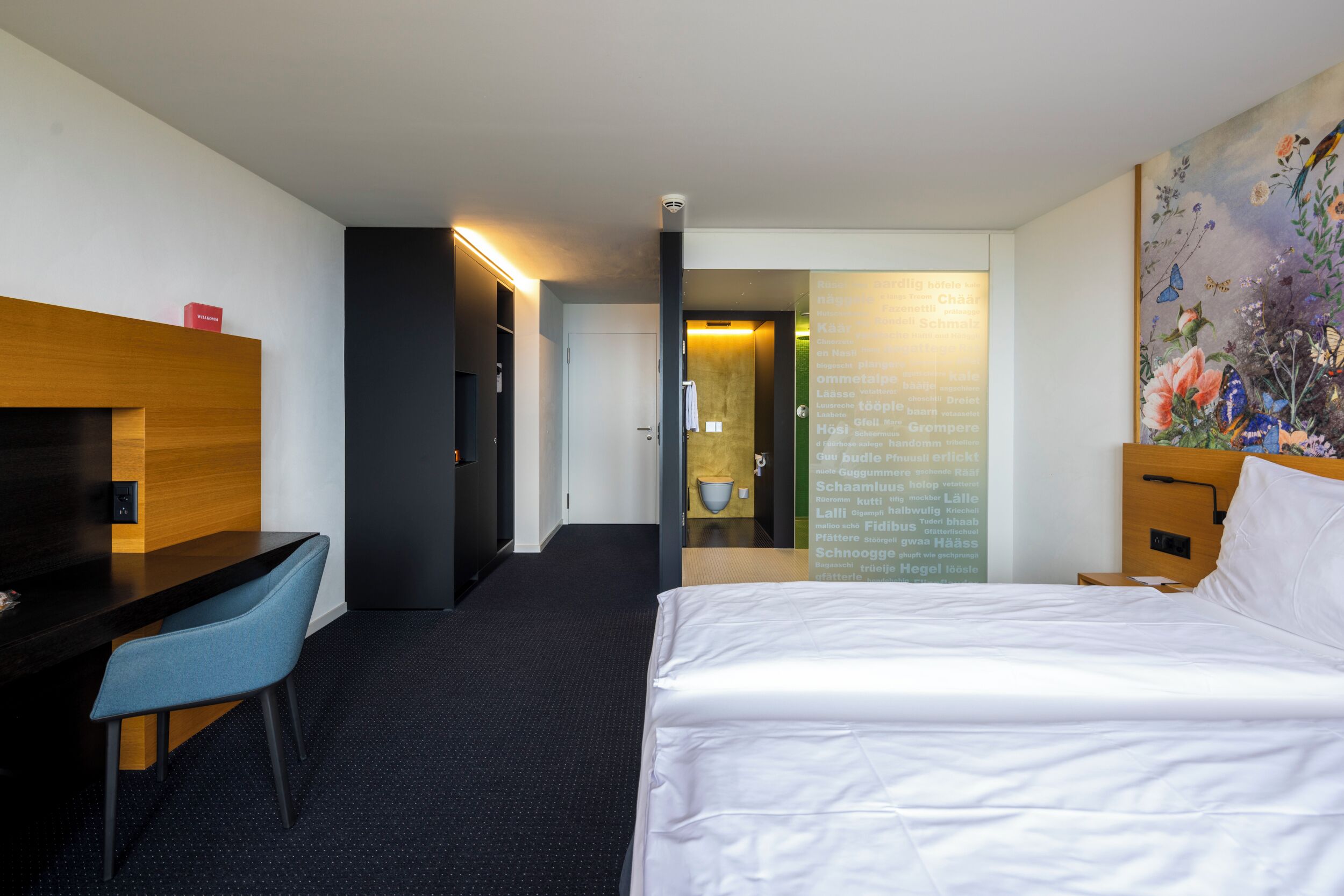 Habitación de hotel con cama, escritorio y silla. Hay un espejo en la pared y un cuadro sobre la cama.