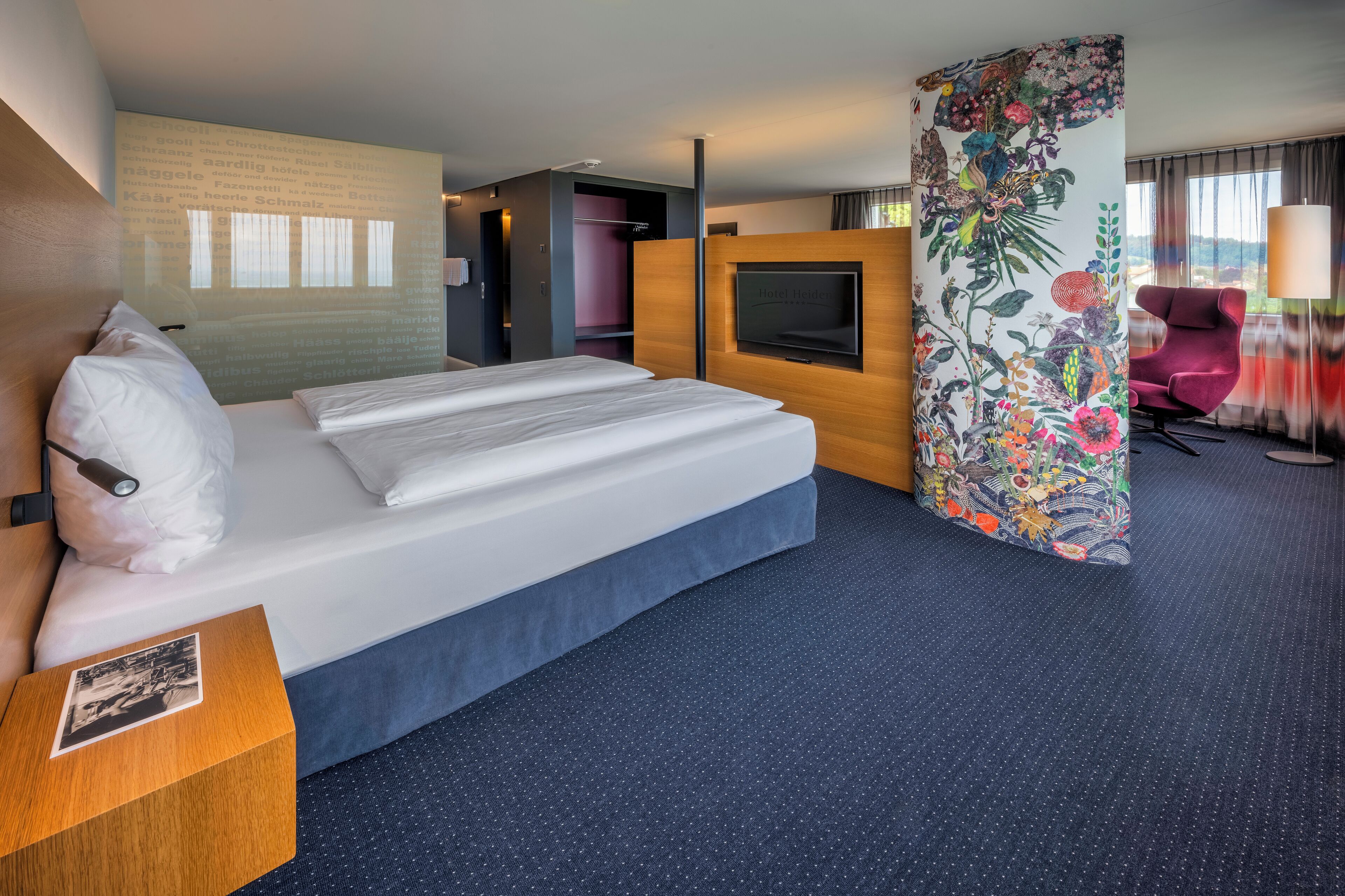 A hotel room with a large bed, a desk with a photo frame, a TV, and a floral-patterned partition.