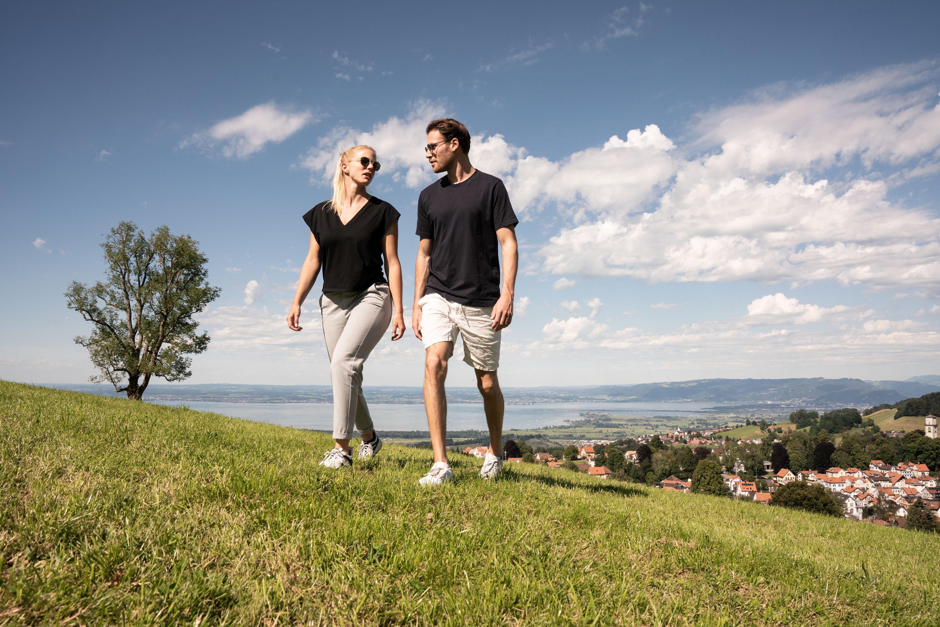 Ein Paar spaziert einen grasbewachsenen Hügel hinauf, im Hintergrund sieht man eine Stadt und einen See.