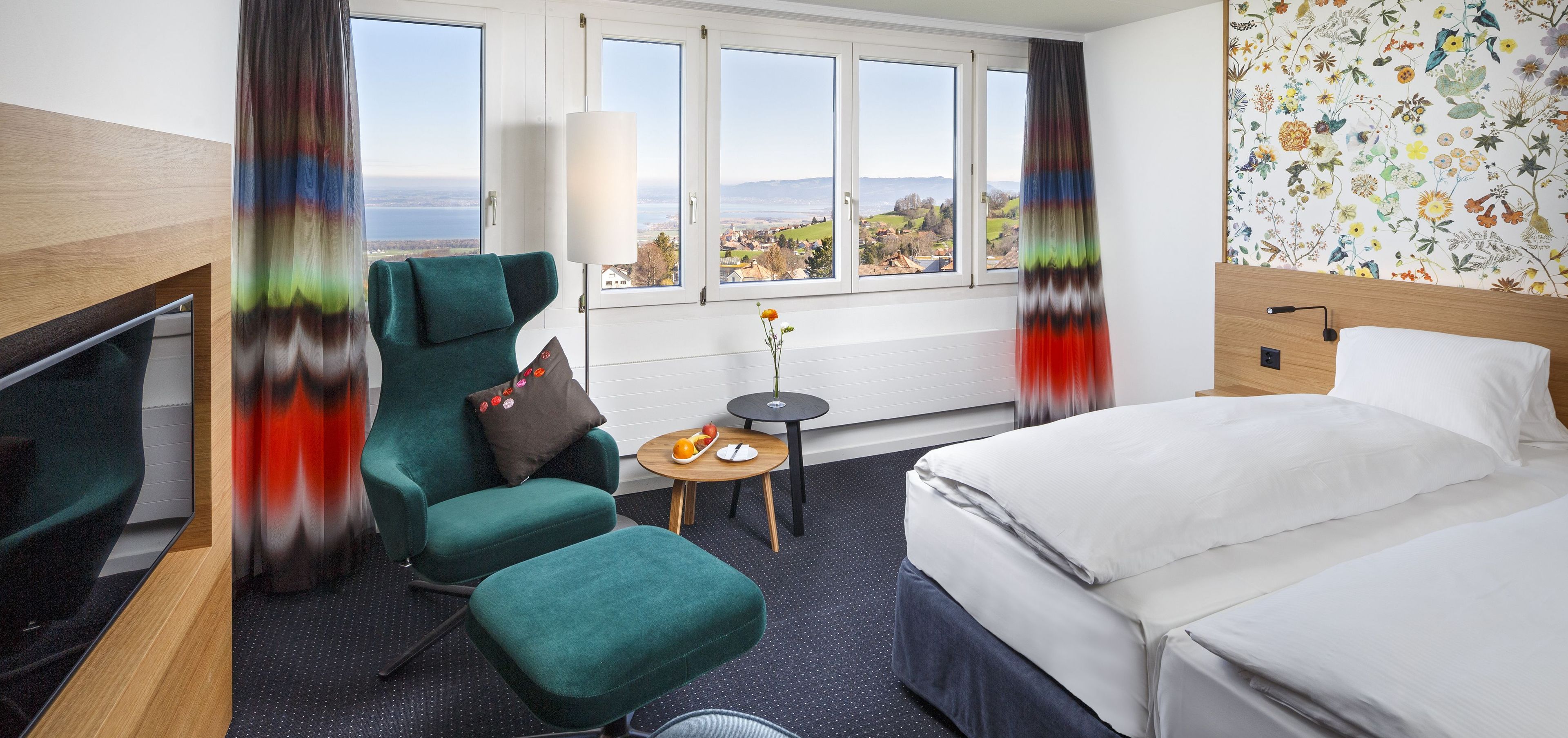 A hotel room with a bed, a desk, a chair, a small table with a fruit bowl, and a view of the landscape through large windows.