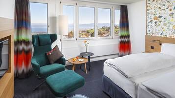 A hotel room with a bed, a desk, a chair, a small table with a fruit bowl, and a view of the landscape through large windows.