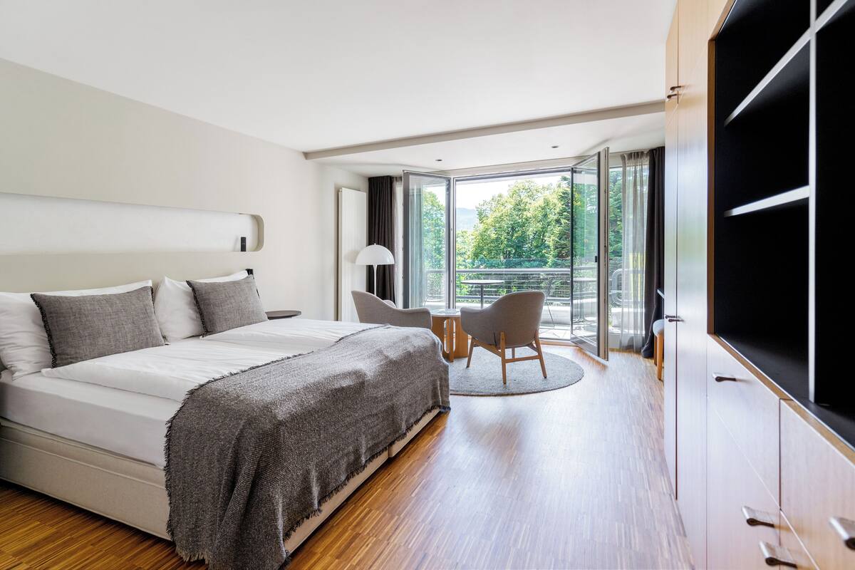 A modern bedroom with a large bed, a desk, and a chair near a sliding glass door leading to a balcony.