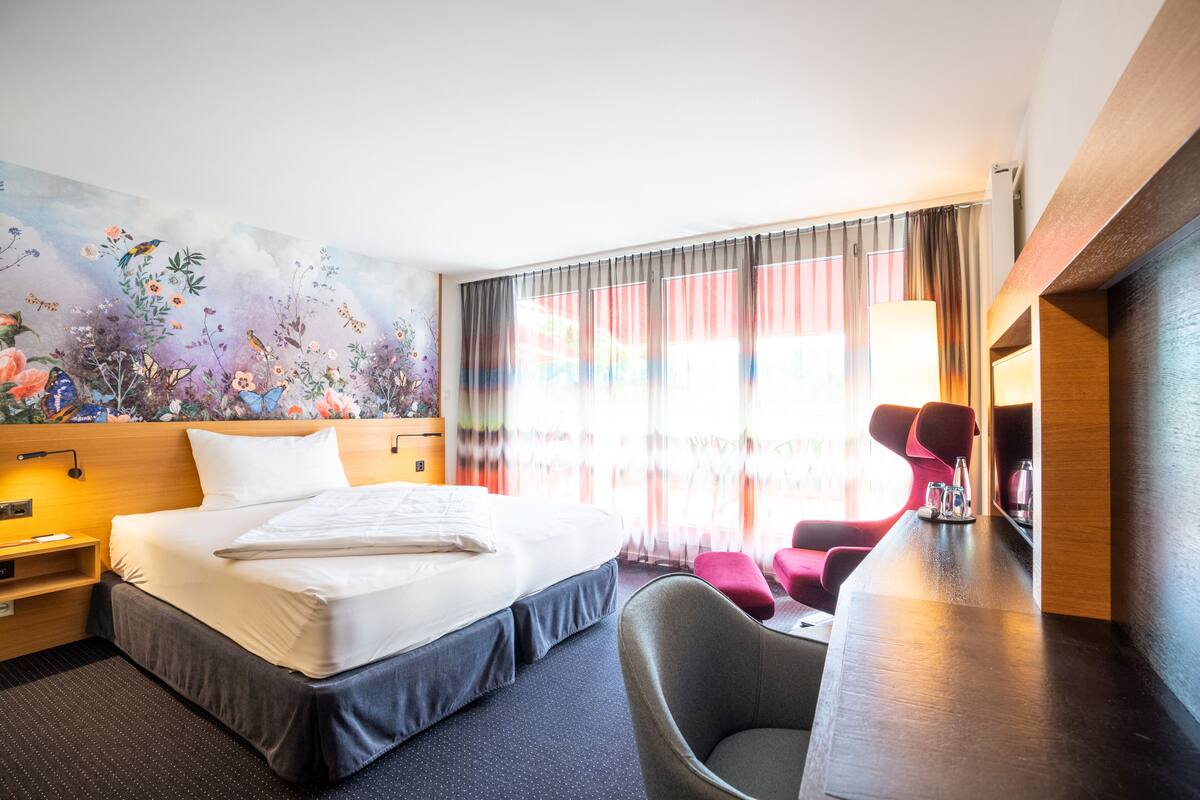 A hotel room with a large bed, a desk, a chair, a nightstand, and a colorful mural on the wall.
