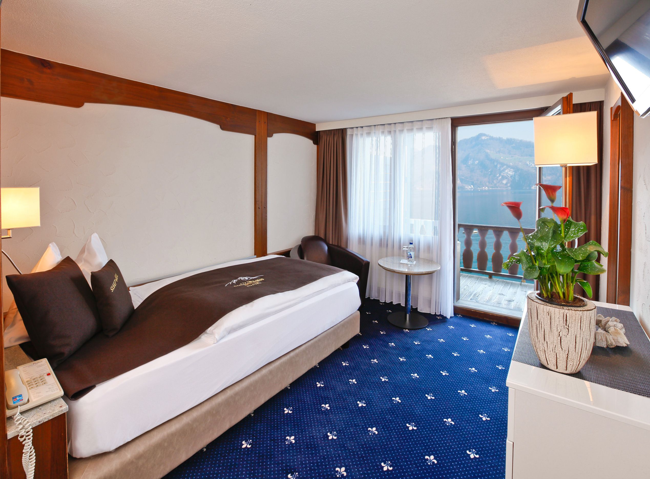A hotel room with a bed, a desk, a chair, a small table, a potted plant, and a view of the outdoors.