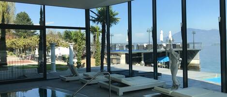 A poolside area with lounge chairs and a statue, large windows offering a view of the water and a distant shoreline.