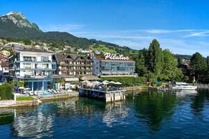 Hotel frente al agua con un llamativo letrero 'Pilatus', un muelle con barcos y montañas al fondo.