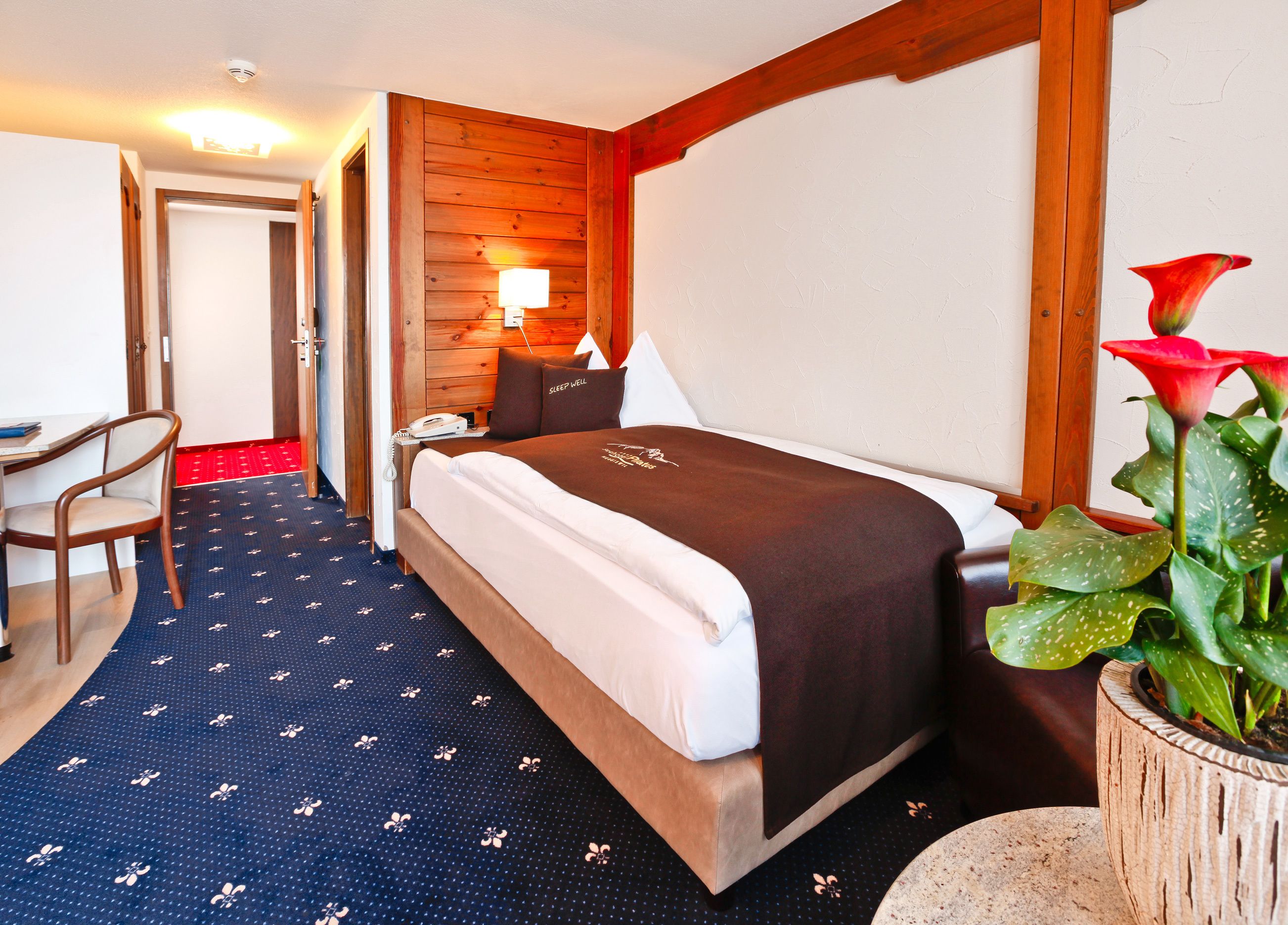 A hotel room with a bed, a desk, a chair, a lamp, and a potted plant.