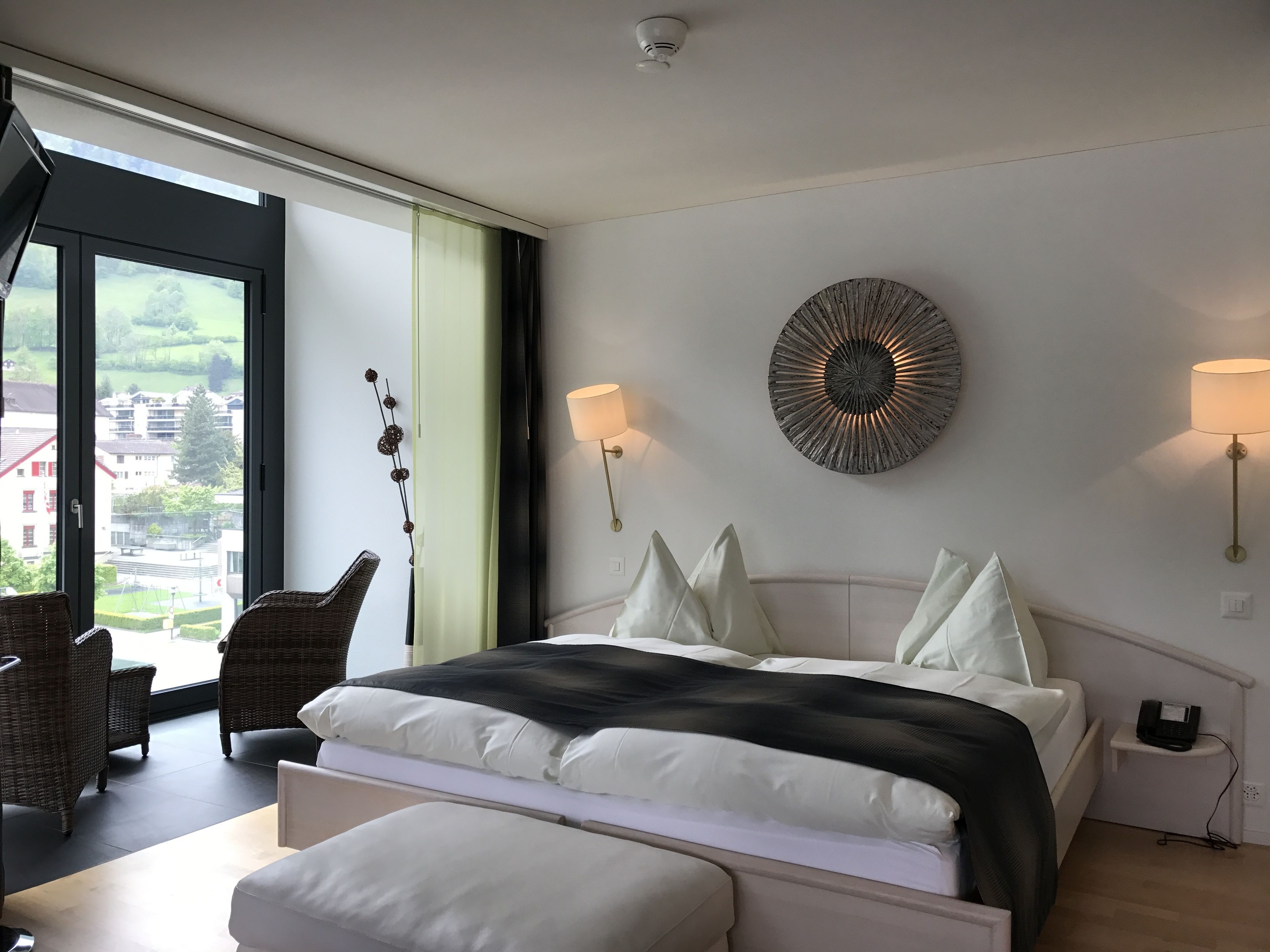A modern bedroom with a large bed, two wall-mounted lamps, a decorative wall piece, and a view of the outside.