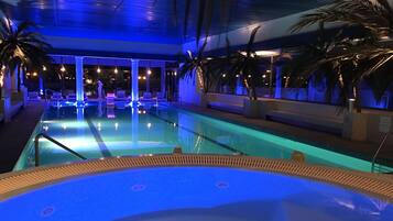 A swimming pool with lounge chairs and palm trees, surrounded by a covered area with blue lighting.