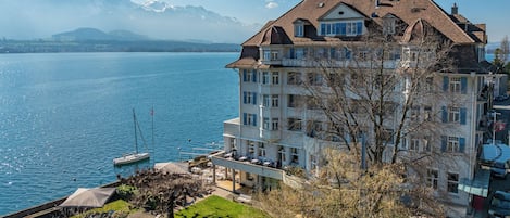 Un hotel de varios pisos con balcones que dan a un lago, un muelle con barcos y montañas al fondo.