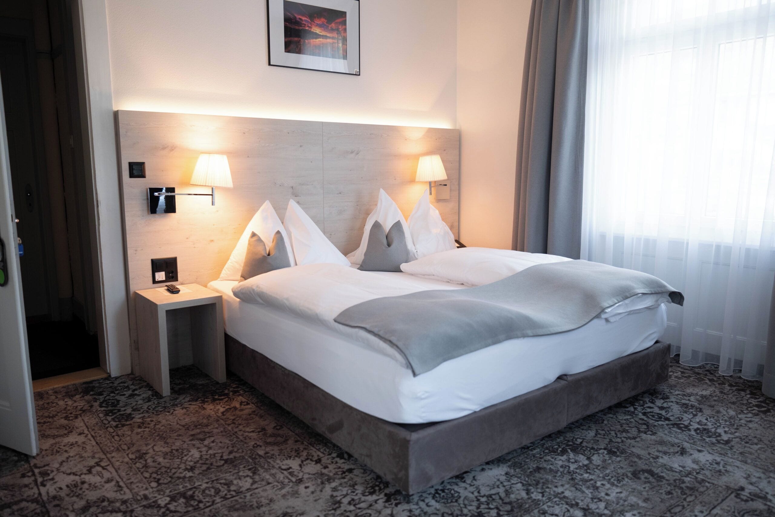 A hotel room with a large bed, two bedside tables, a wall-mounted lamp, and a framed picture above the bed.