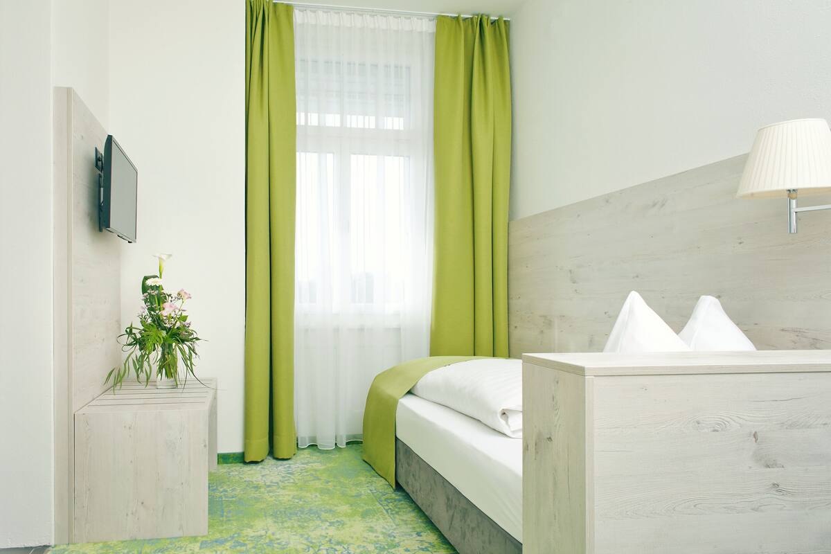 A hotel room with a bed, a bedside table with a lamp, a TV mounted on the wall, and a window with green curtains.