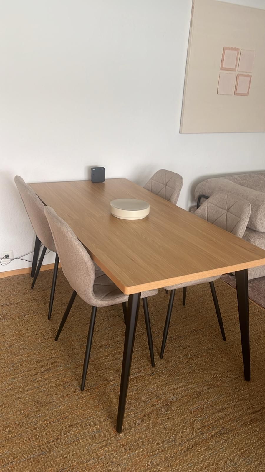 A rectangular wooden dining table with four chairs, a small gray object on the table, and a beige couch in the background.