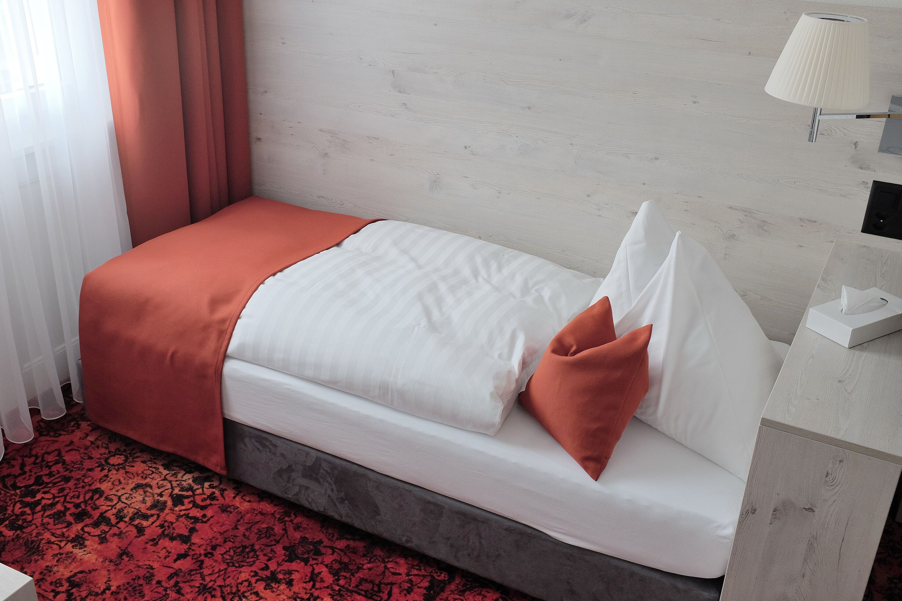 Una cama individual con colchón blanco, manta de color naranja y almohadas, alfombra roja estampada y una lámpara blanca sobre una mesita de noche.