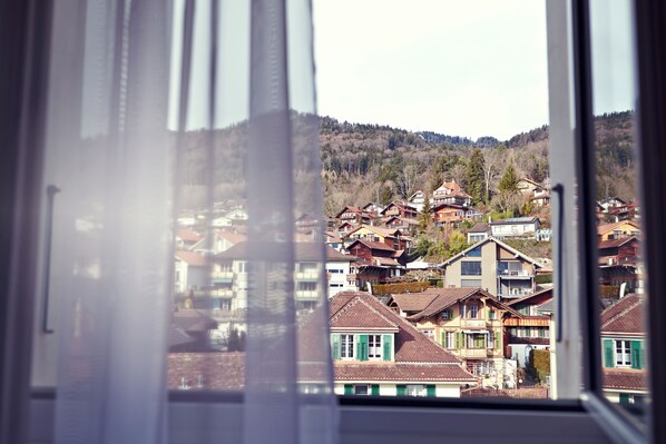 Blick durch ein Fenster auf ein Wohngebiet mit Häusern, einige davon mit Balkonen, und eine hügelige Landschaft im Hintergrund.