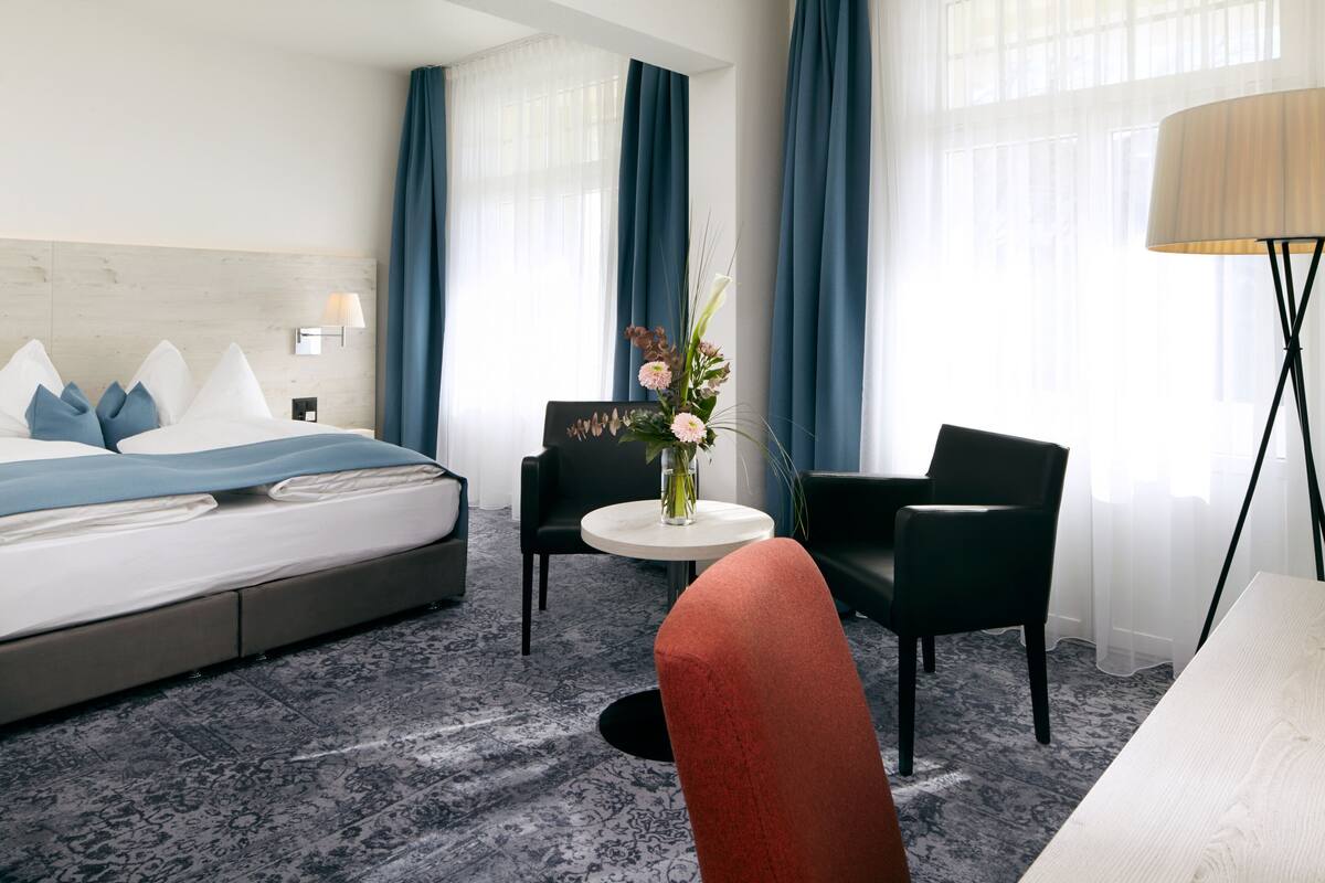 A hotel room with a bed, a round table with chairs, a vase with flowers, and a lamp.
