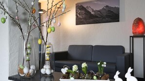 A modern living room with a black sofa, a glass vase with branches and eggs, a framed mountain landscape, and a red crystal on a stand. - Crystal Interlaken (Interlaken)