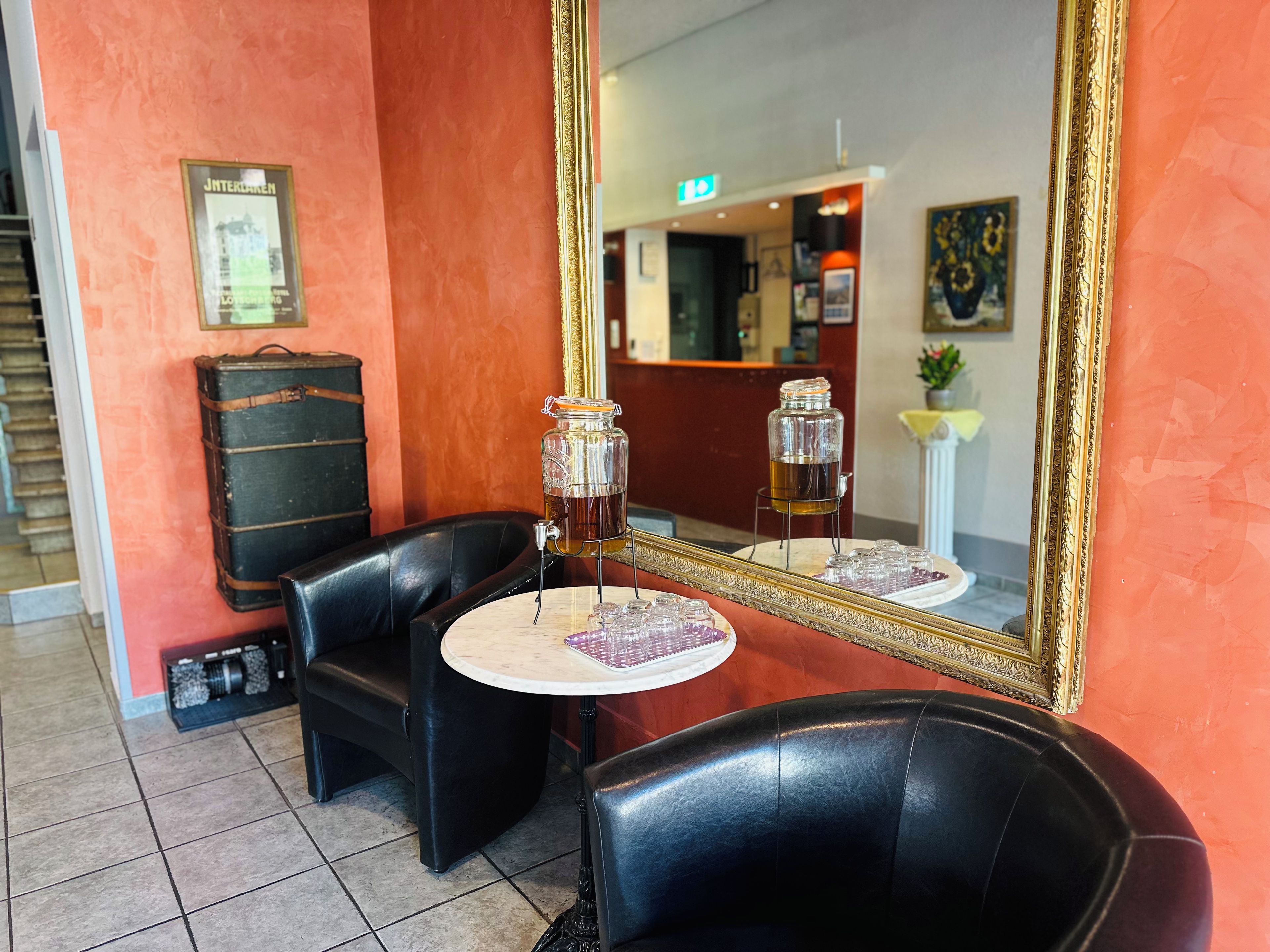 Een kamer met een grote spiegel, een tafel met een glazen pot, een zwarte lederen stoel en een koffer tegen een rode muur.