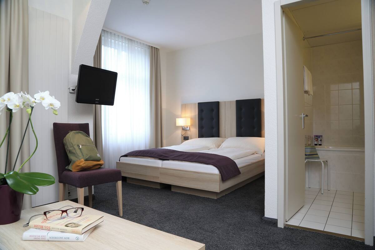 Una habitación de hotel con una cama, un televisor montado en la pared, un escritorio con una planta y libros, una silla y un rincón de ducha.