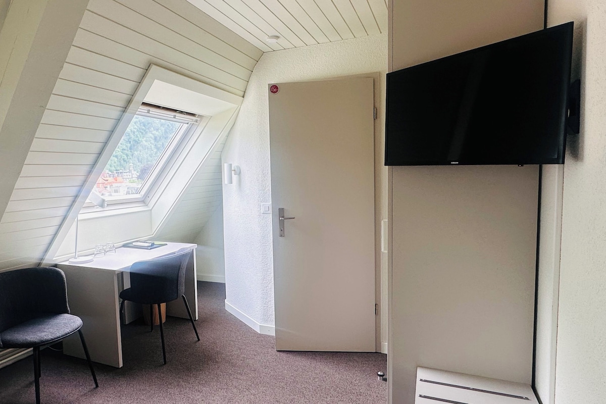 Una habitación pequeña e inclinada con un escritorio, dos sillas, un televisor y una puerta.