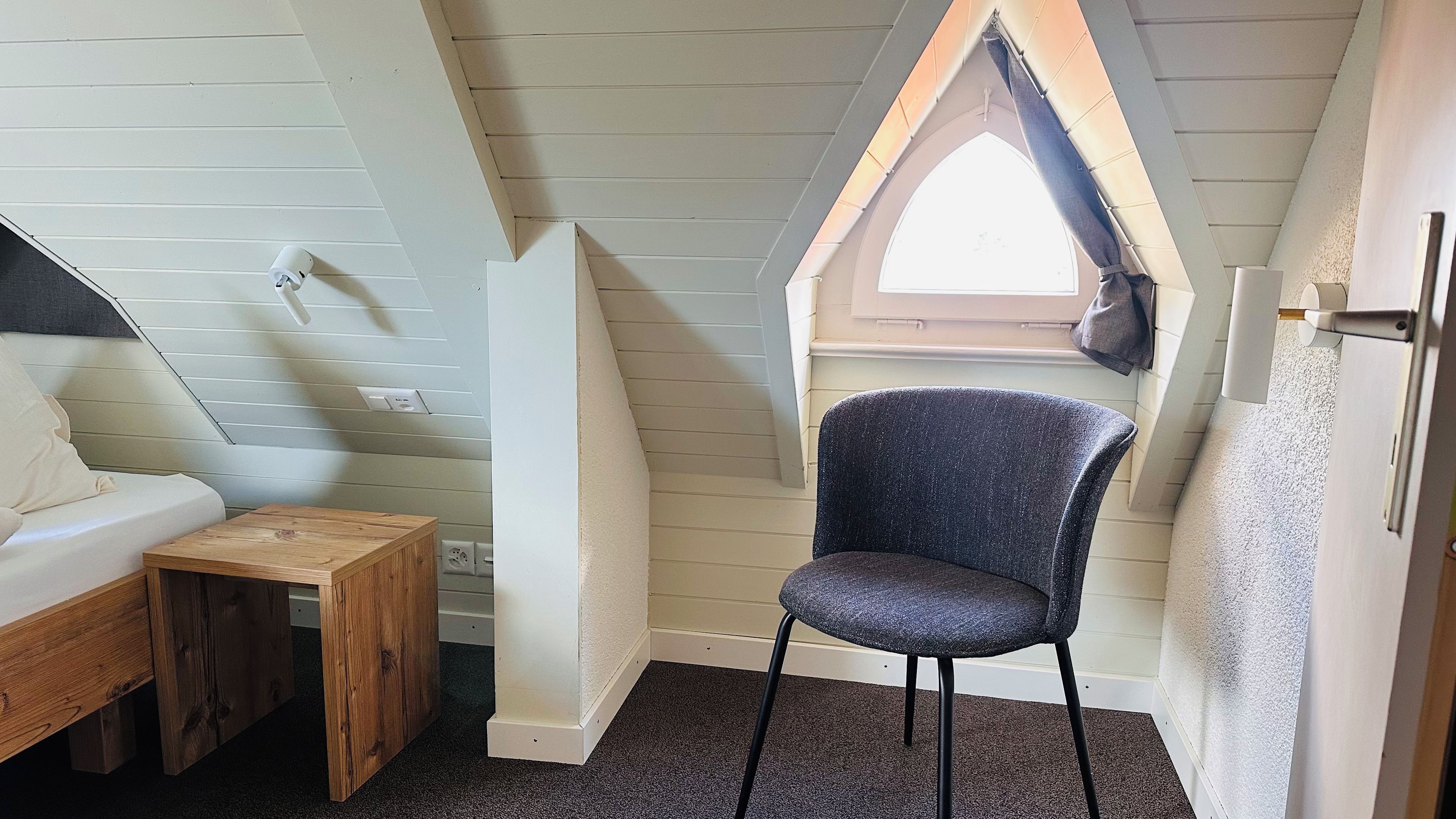 A small, cozy room with a sloped ceiling, a window with a curtain, a wooden table, and a chair.