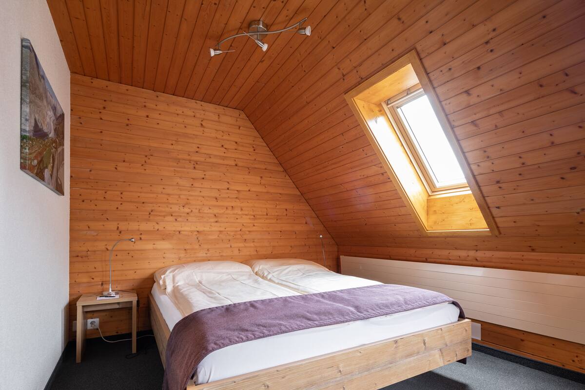 Un dormitorio con techo de madera inclinado, un tragaluz, una cama con manta púrpura y una mesita de noche con lámpara.