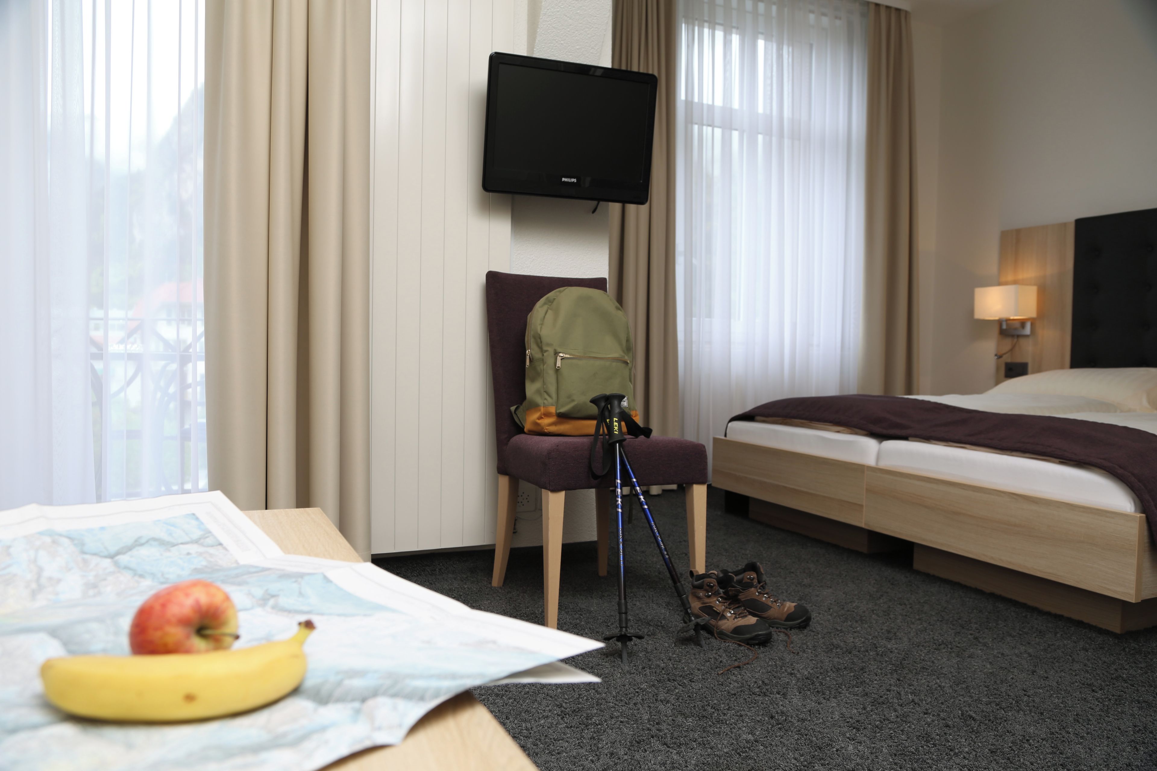 Una habitación de hotel con una cama, un escritorio con una banana y una manzana, una silla con una mochila, un trípode y un televisor en la pared.