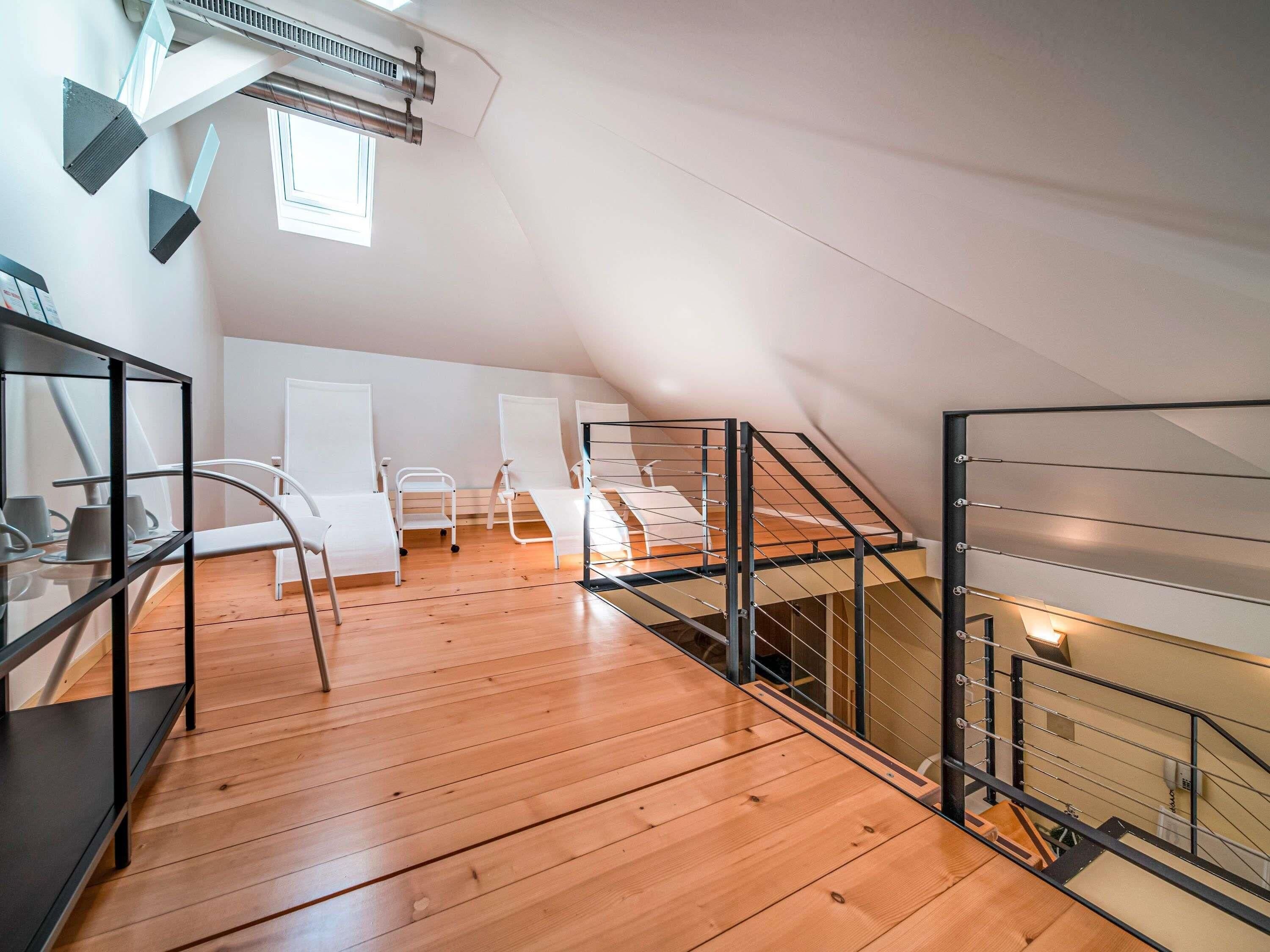 Ein Dachzimmer mit Holzboden, weißen Stühlen, einem Glastisch und einer Metalltreppe.