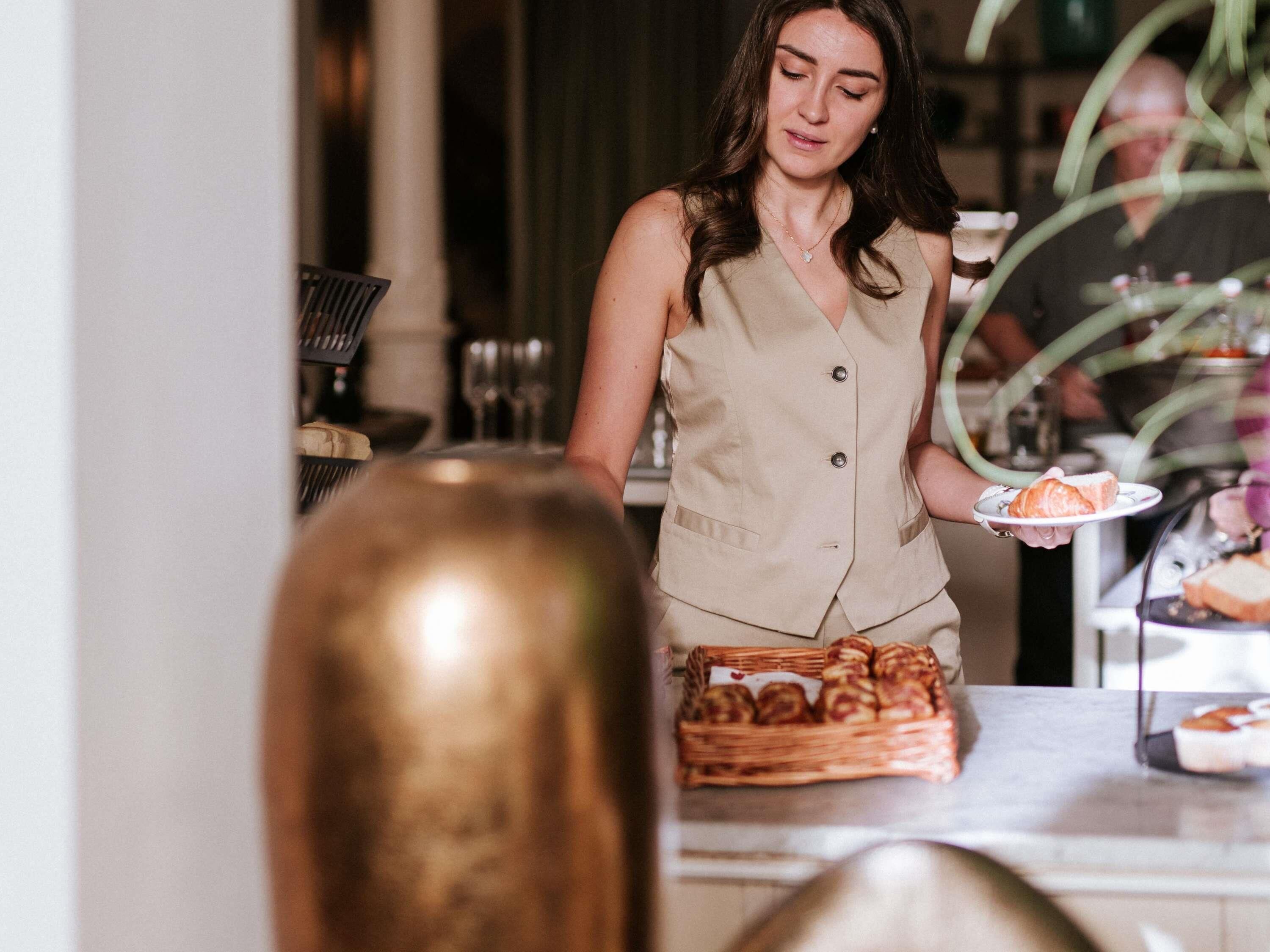 A person serving food at a table with a basket of pastries and a plate of food.