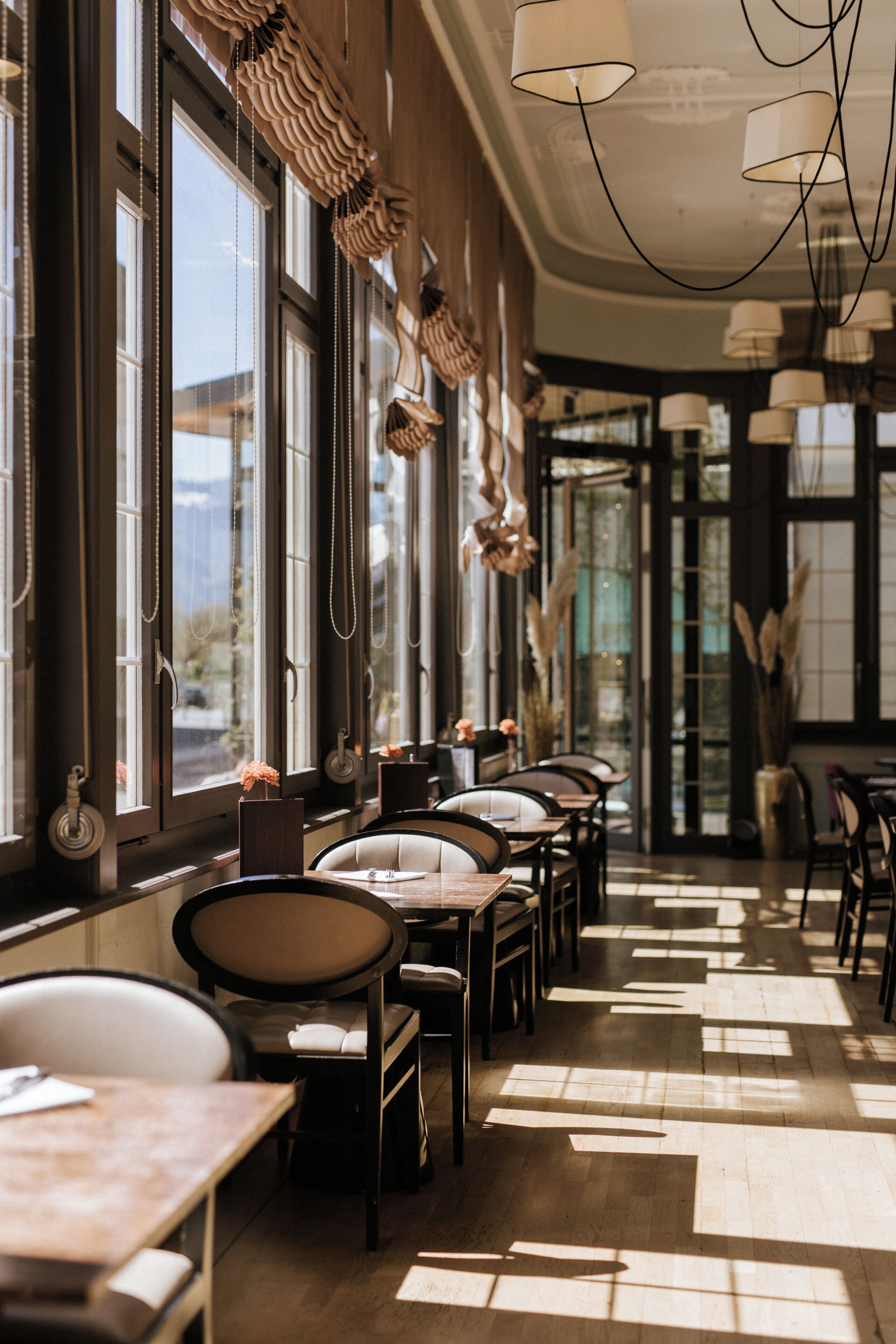 A restaurant interior with long tables, chairs, hanging light fixtures, and large windows allowing natural light.