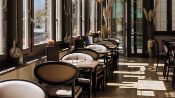 A restaurant interior with long tables, chairs, hanging light fixtures, and large windows allowing natural light.