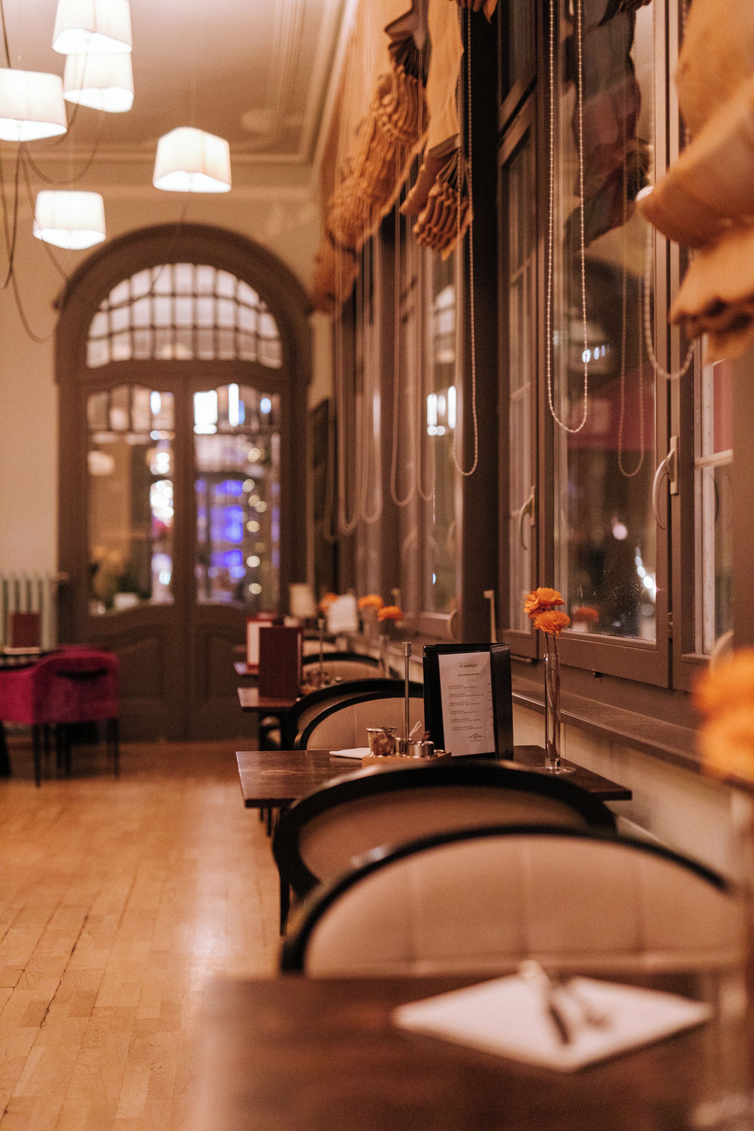 A restaurant interior with wooden tables, chairs, hanging lights, and a large arched doorway.