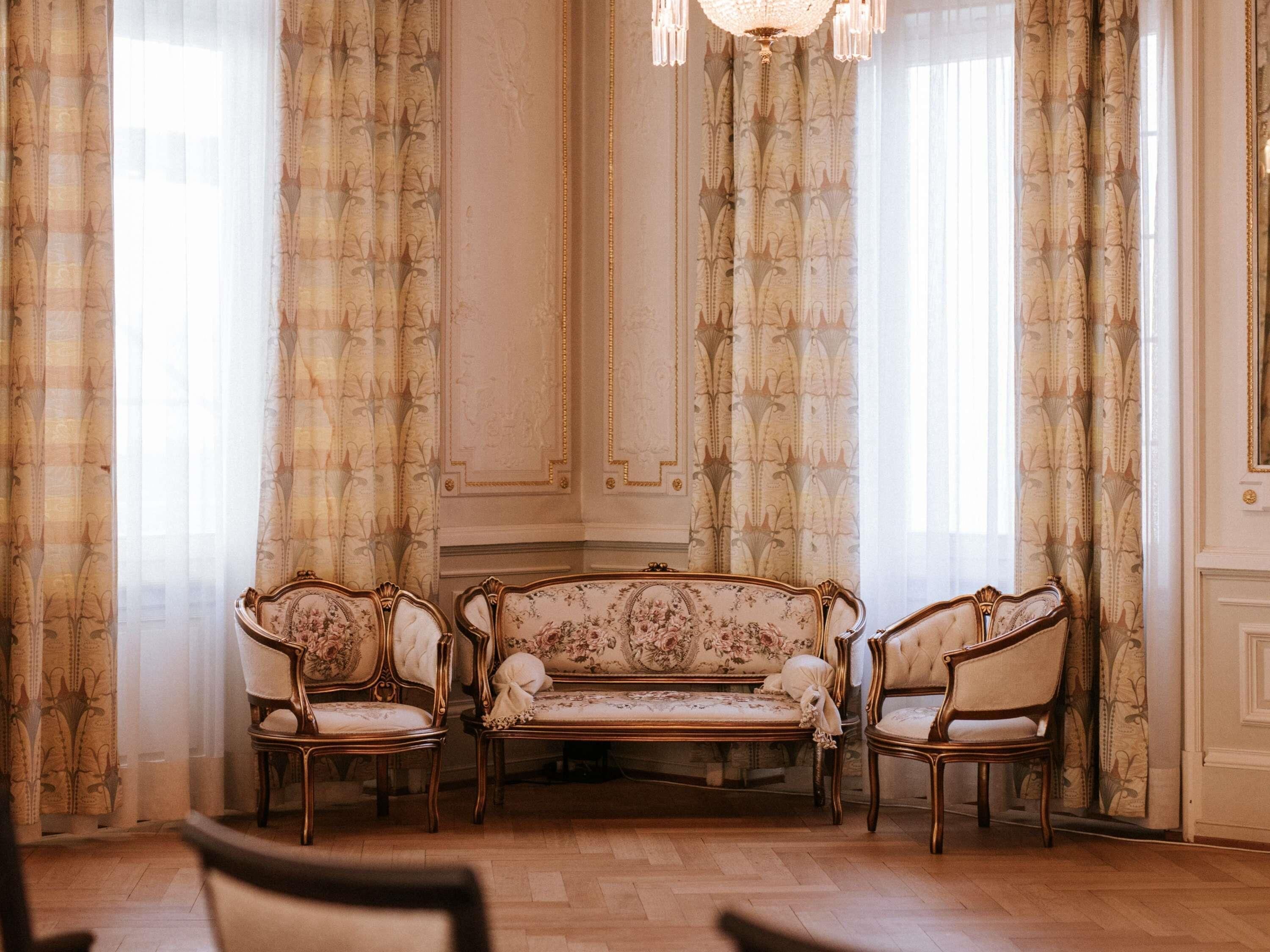 A room with a sofa set, two chairs, a chandelier, and patterned curtains.