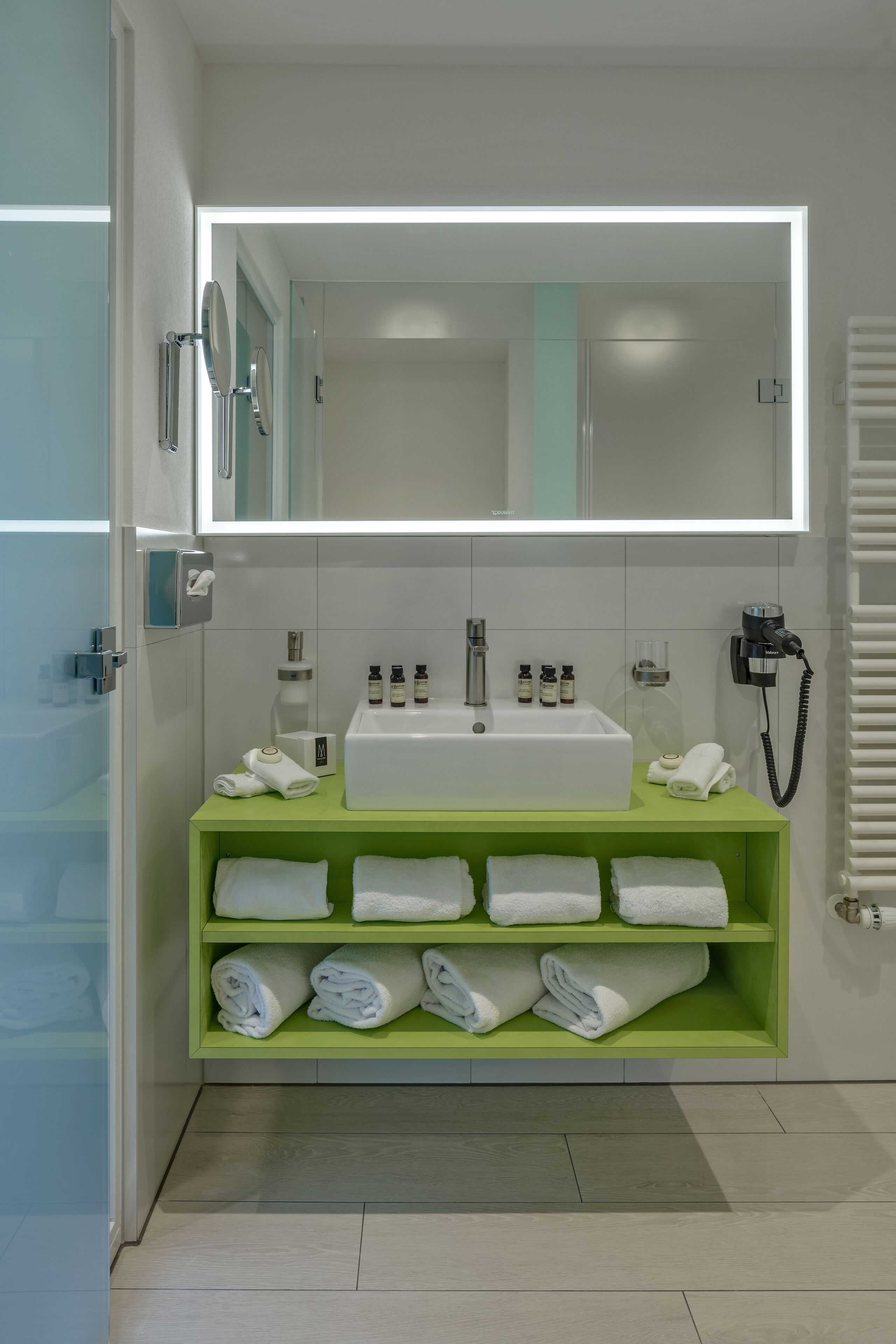 A modern bathroom with a white sink, green shelves, and a large mirror.