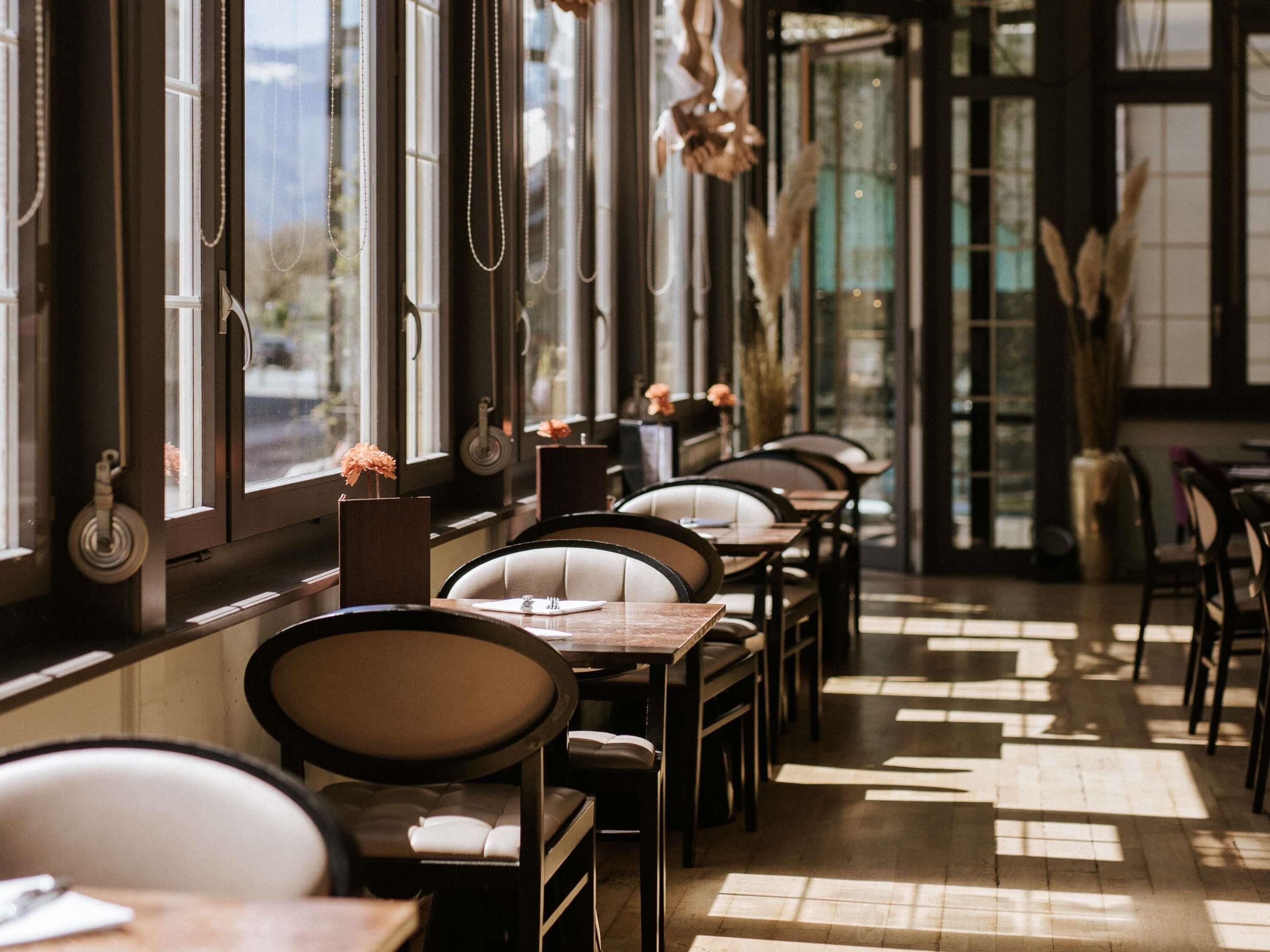 Un restaurante con mesas y sillas dispuestas a lo largo de una pared con ventanas, con luz solar entrando.