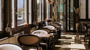 Un restaurante con mesas y sillas dispuestas a lo largo de una pared con ventanas, con luz solar entrando.