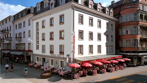Un hotel con varios pisos, mesas al aire libre con sombrillas rojas y un cartel que indica el nombre del hotel.