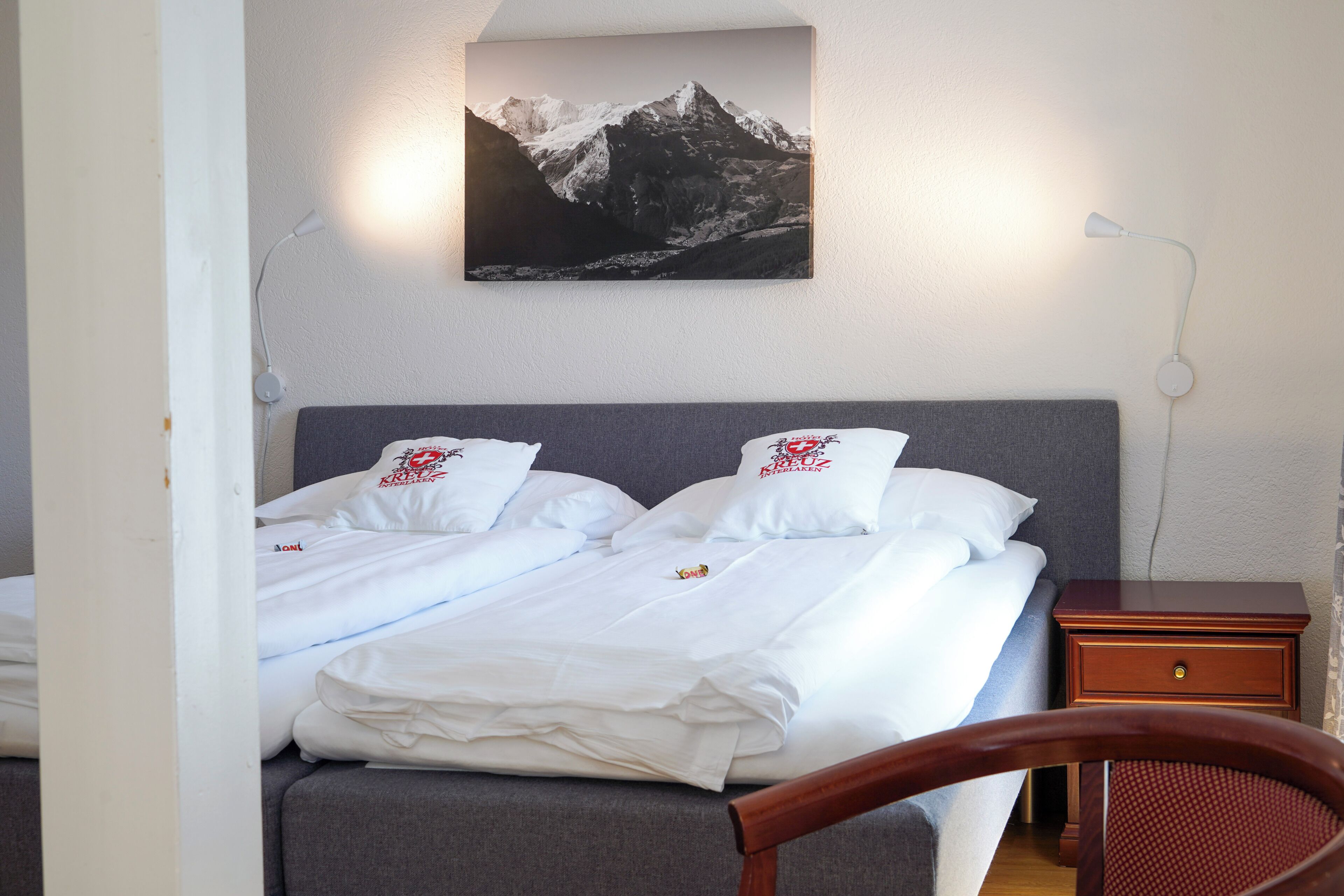 A neatly made bed with white linens and pillows, a gray headboard, and a mountain landscape painting above it.